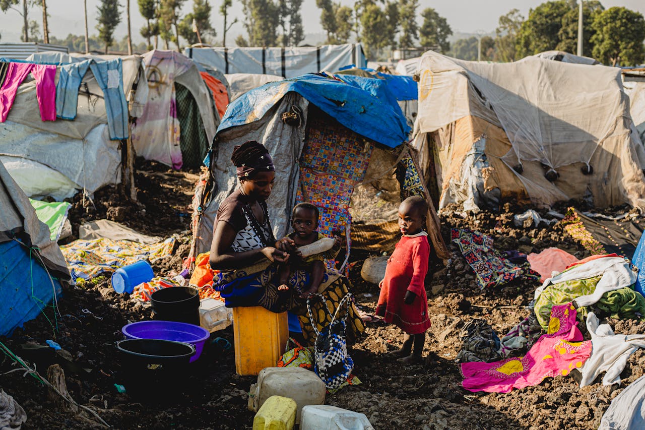 gallery-1 Black woman with children in a makeshift camp setting amidst tents and scattered items.